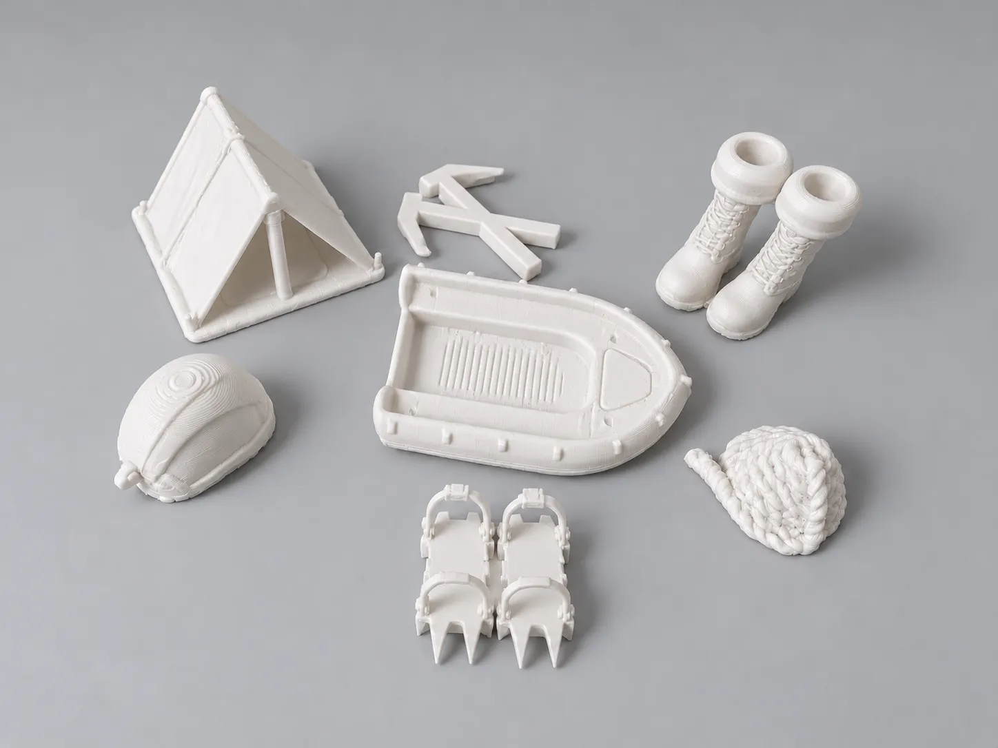 The 3D-printed equipment pieces in white on a grey table: a tent, a crossed ice axe, a pair of snow boots, an inflatable boat, a helmet, a coiled rope and a pair of crampons.