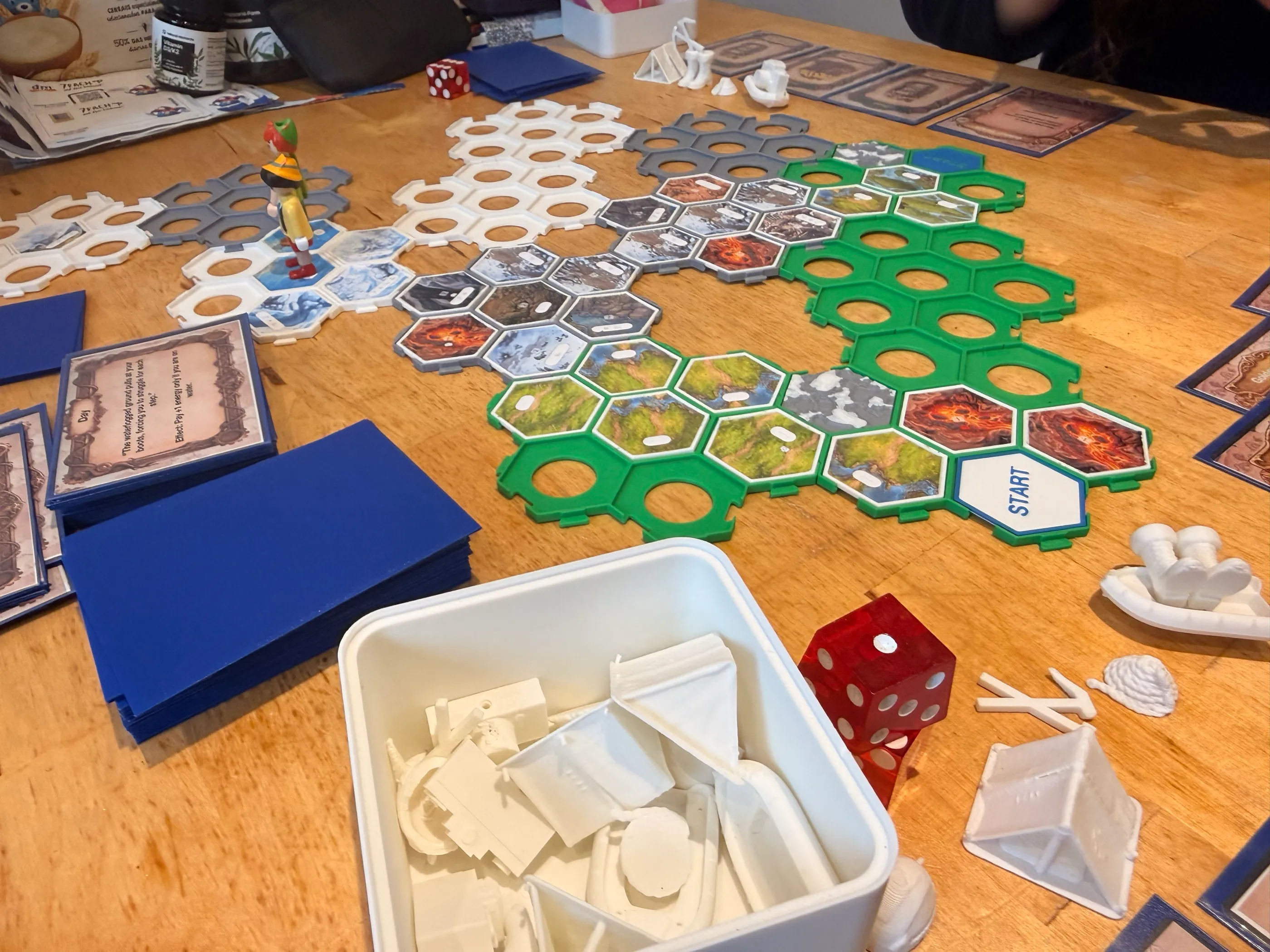 A game in progress: green, grey and white hexagonal frames on a wooden table, partly filled with meadow, rock, snow and lava tiles, a START tile at the foot and a climber figure near the top.